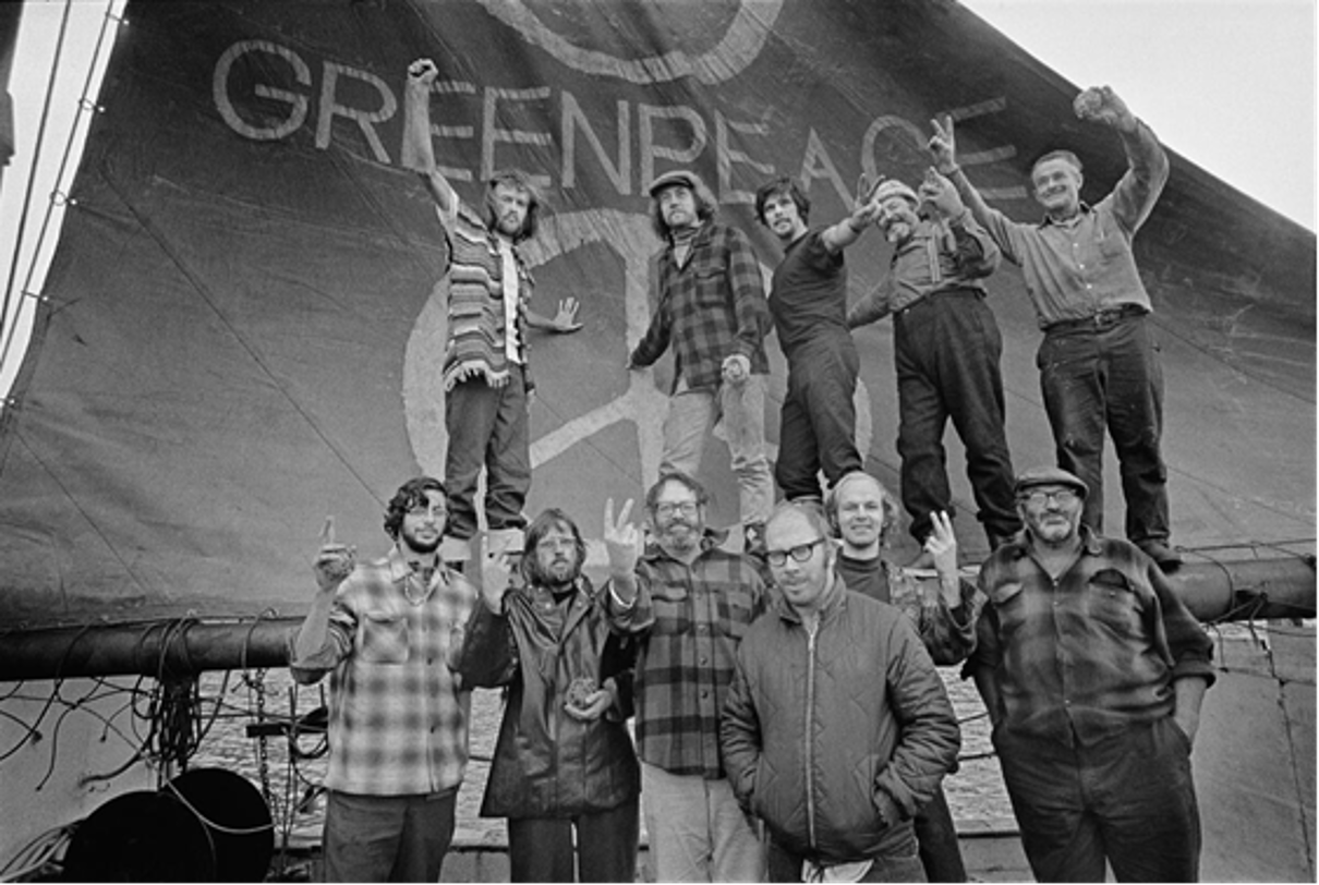 eine Gruppe von Männern vor einem Segelmast, auf dem „Greenpeace“ steht. 