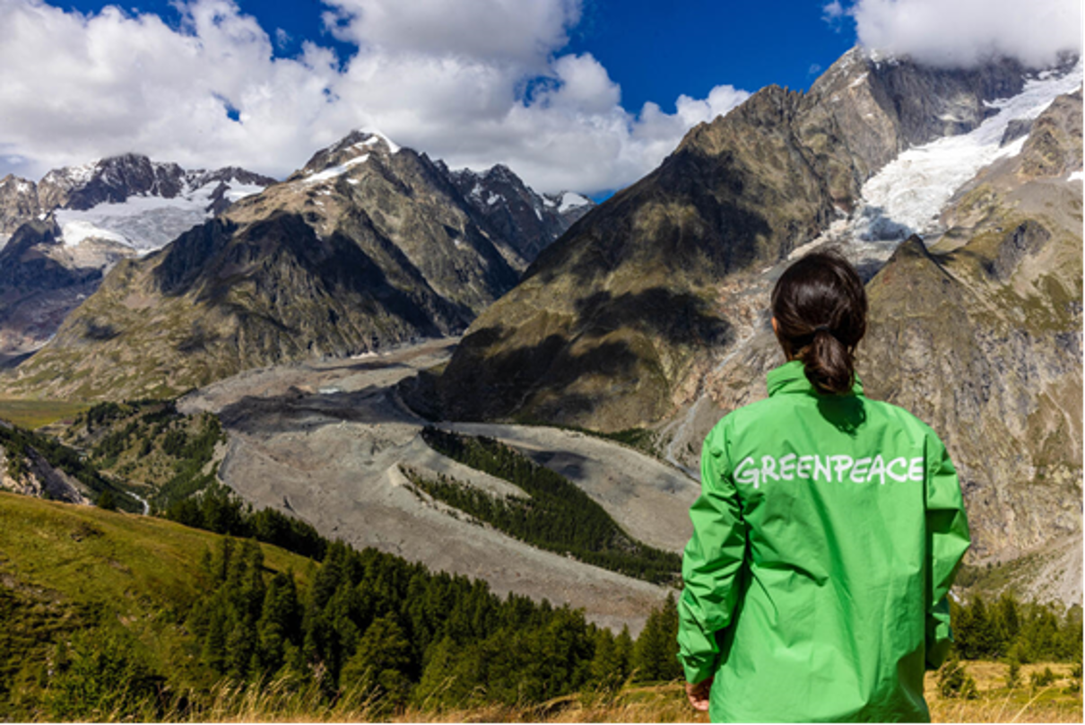 Eine Frau in einer grünen Jacke, auf der „Greenpeace“ steht vor einem Bergpanorama. 