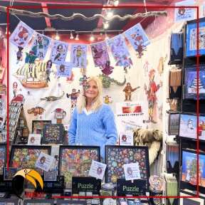 Eine blondhaarige Frau an einem buntgeschmückten Messestand. 