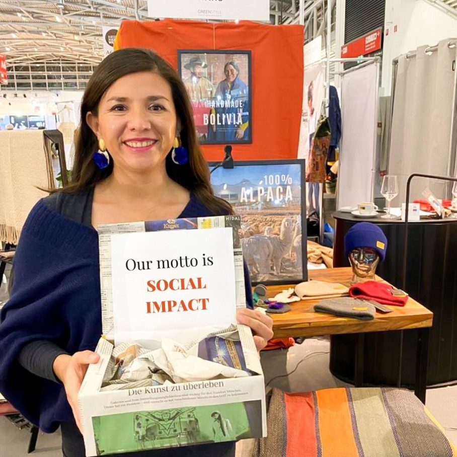 Eine Frau steht an einem Ausstellungsstand und hält eine Box mit der Aufschrift „Our motto is Social Impact“ in den Händen. 