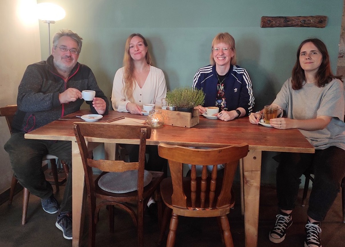 Vier Personen an einem Tisch sitzend, alle lächeln und blicken in die Kamera. 